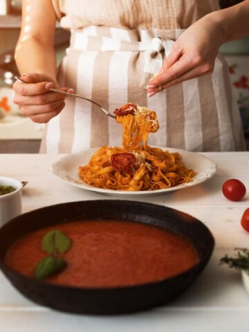 Young woman in kitchen making delicious pasta with sauce using a pan at home. Italian rural cooking still life. Wooden board, fresh vegetables, cooked pasta and a pan of sauce on wooden kitchen table.