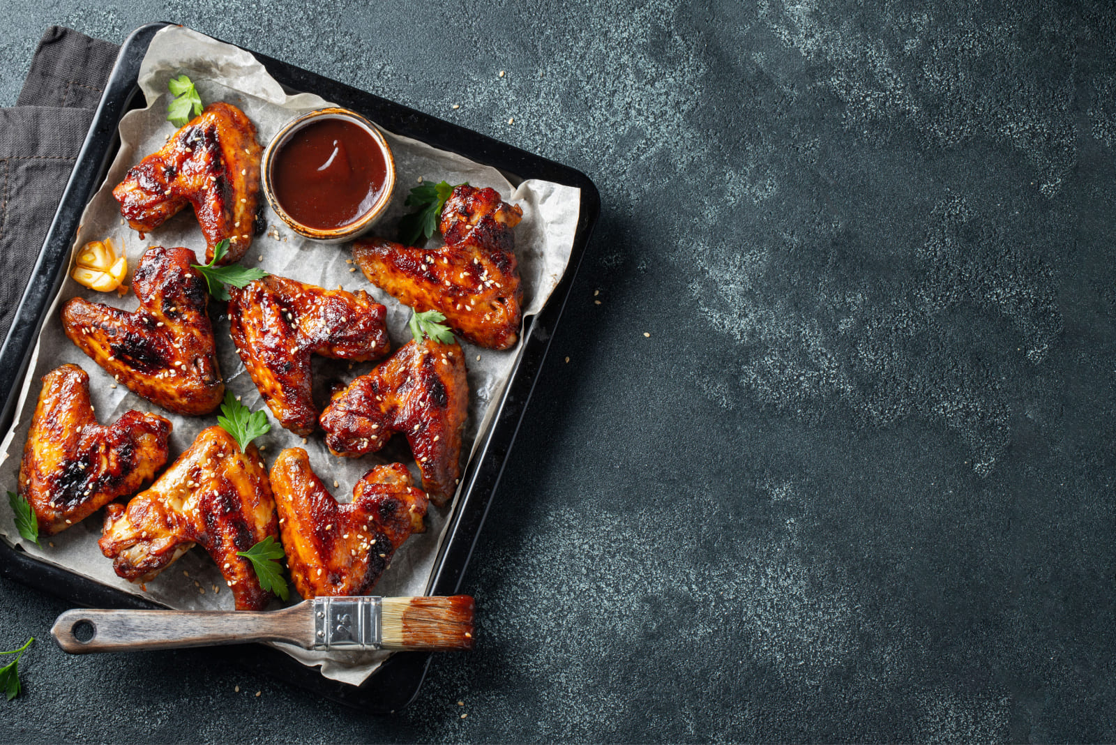 Roasted chicken wings in barbecue sauce with sesame seeds and parsley in a baking tray on a dark table. Top view with copy space. Tasty snack for beer on a dark background. Flat lay.
