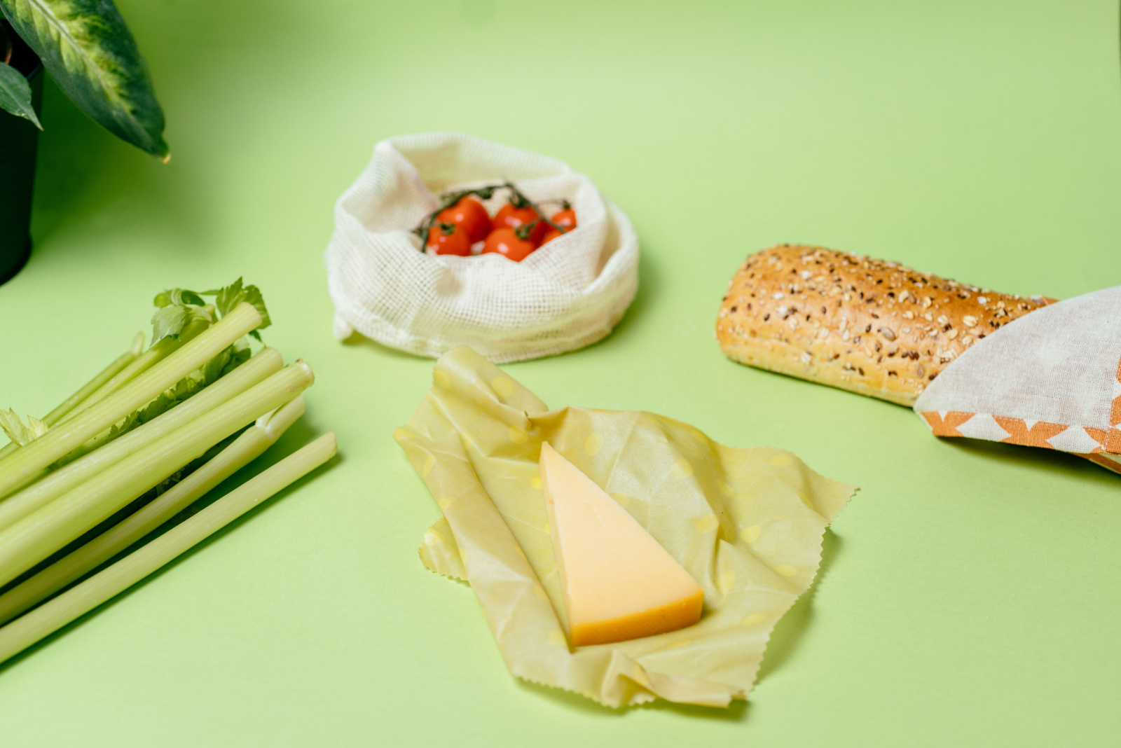 Cheese In Reusable Environmentally Friendly Beeswax Wrap. Cherry tomatoes in cotton bag, celery, baguette wrapped in kitchen textile on green background. Plactic free, zero waste concept.