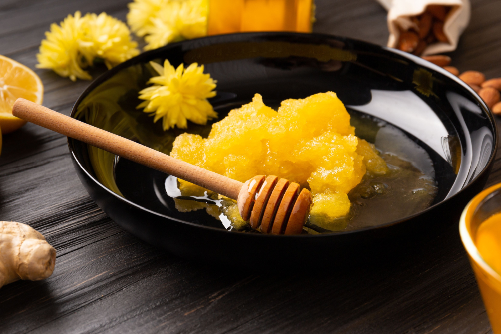 Crystallized honey with dipper in a black round plate and yellow flowers on a dark wooden background.
