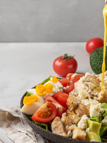 A hand pouring salad dressing into cobb salad with chicken, avocado, bacon, blue cheese, tomato and eggs. Keto diet food. Classic American dish