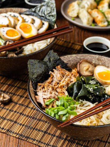 Traditional ramen with jerked pork or chicken. With udon or ramen noodles. Served in classic bowls. Gyoza dumplings and mushrooms in the background. Natural wooden background.