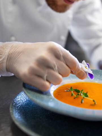 food cooking, profession and people concept - close up of male chef with flower serving plate of pumpkin cream soup at restaurant kitchen