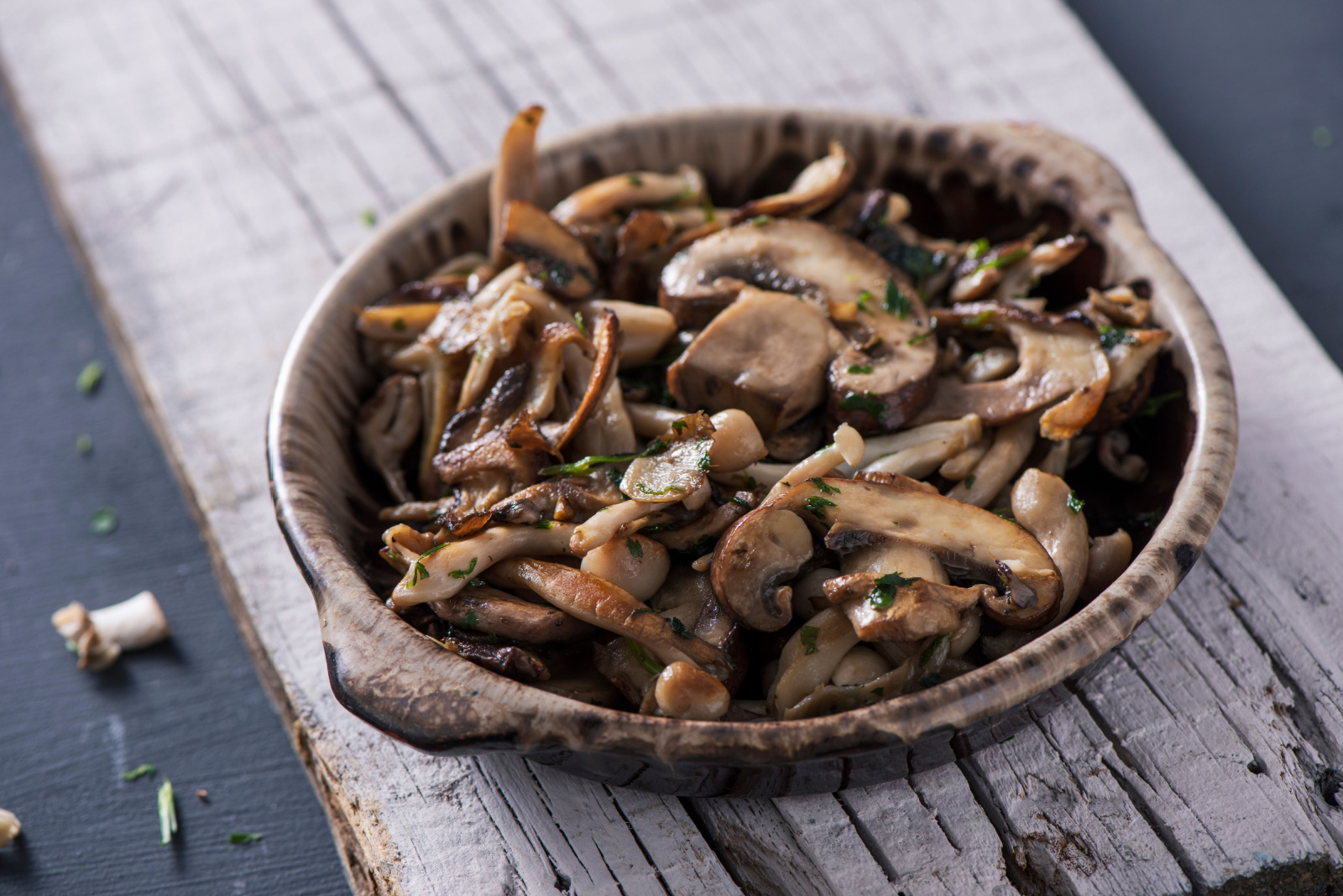 closeup of a brown earthenware bowl with some cooked mixed mushrooms, such as common mushrooms, oyster mushrooms or shiitake, on a white rustic wooden board