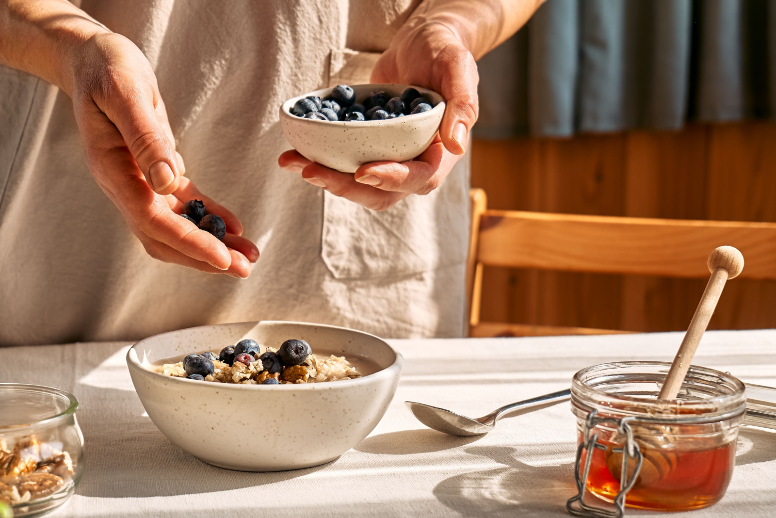 Woman preparing healthy dieting vegan nutritious breakfast. Female hand putting blueberries in the bowl with oatmeal porridge with walnuts and honey.